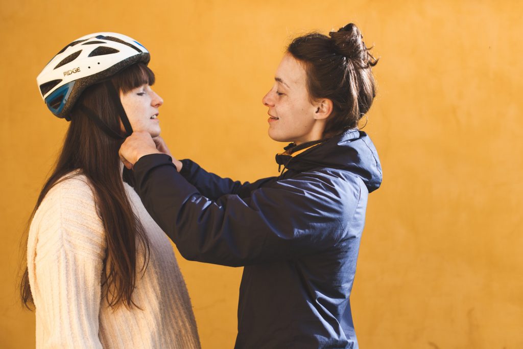 A women with brown hair in a bun secures a helmet on a person with long brown hair